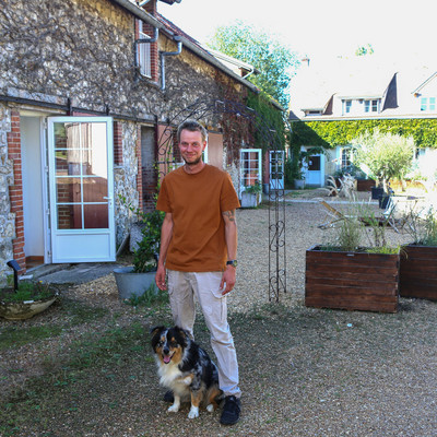 Homme debout avec un chien assis entre ses jambes, devant une charmante maison