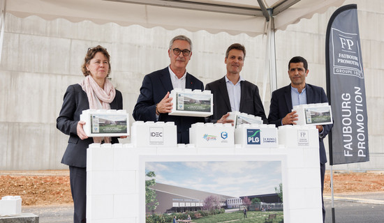 Quatre personnes tenant des blocs symboliques lors d'une cérémonie officielle sous une tente, devant une maquette représentant un projet de construction, avec des logos d'entreprises partenaires en arrière-plan